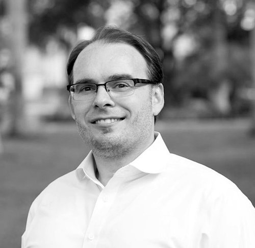 Gavin Hurley. Black and white headshot. Gavin's wearing a crisp white shirt. He has two-day stubble, is wearing scholarly rectangle glasses and has short hair parted to the left. 