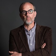 Portrait of Michael Kramp. Caucasian male with grey stubble. Balding with round dark eyeglasses. He stands armed crossed in a dark blazer and checked shirt looking straight at the camera with a serious expression.